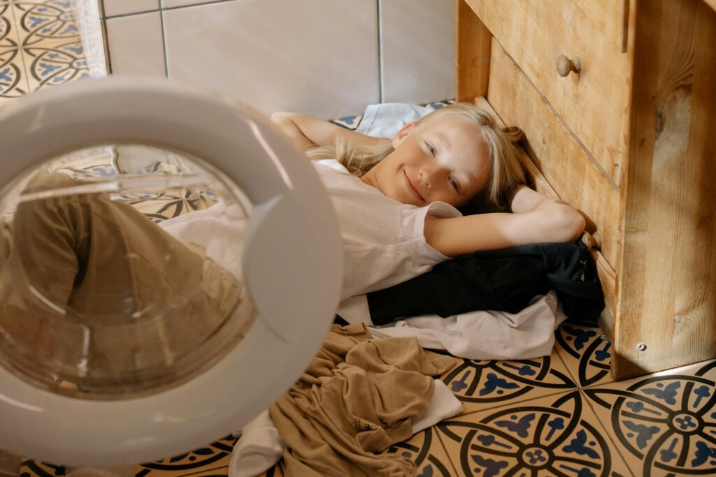 A smiling young girl lies on a tiled floor surrounded by laundry indoors.
